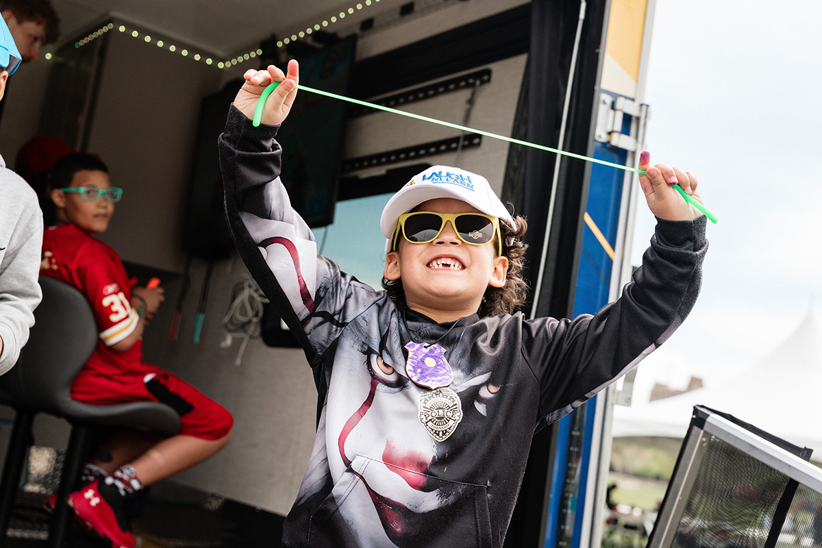 Child wearing a clown face hoodie and a police badge, holding a green string stretched above their head.