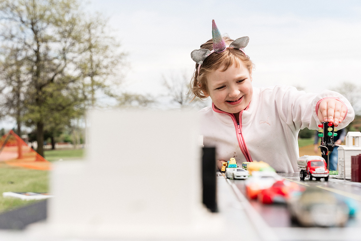 Child wearing a unicorn headband playing with toy cars and a traffic light on a miniature road setup outdoors.
