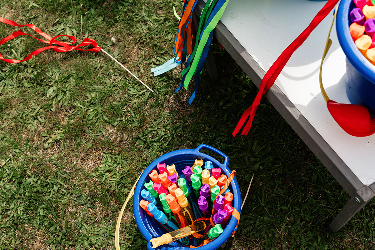 Blue bucket filled with colorful bubble wands on grass next to a white table with another bucket and red ribbons.