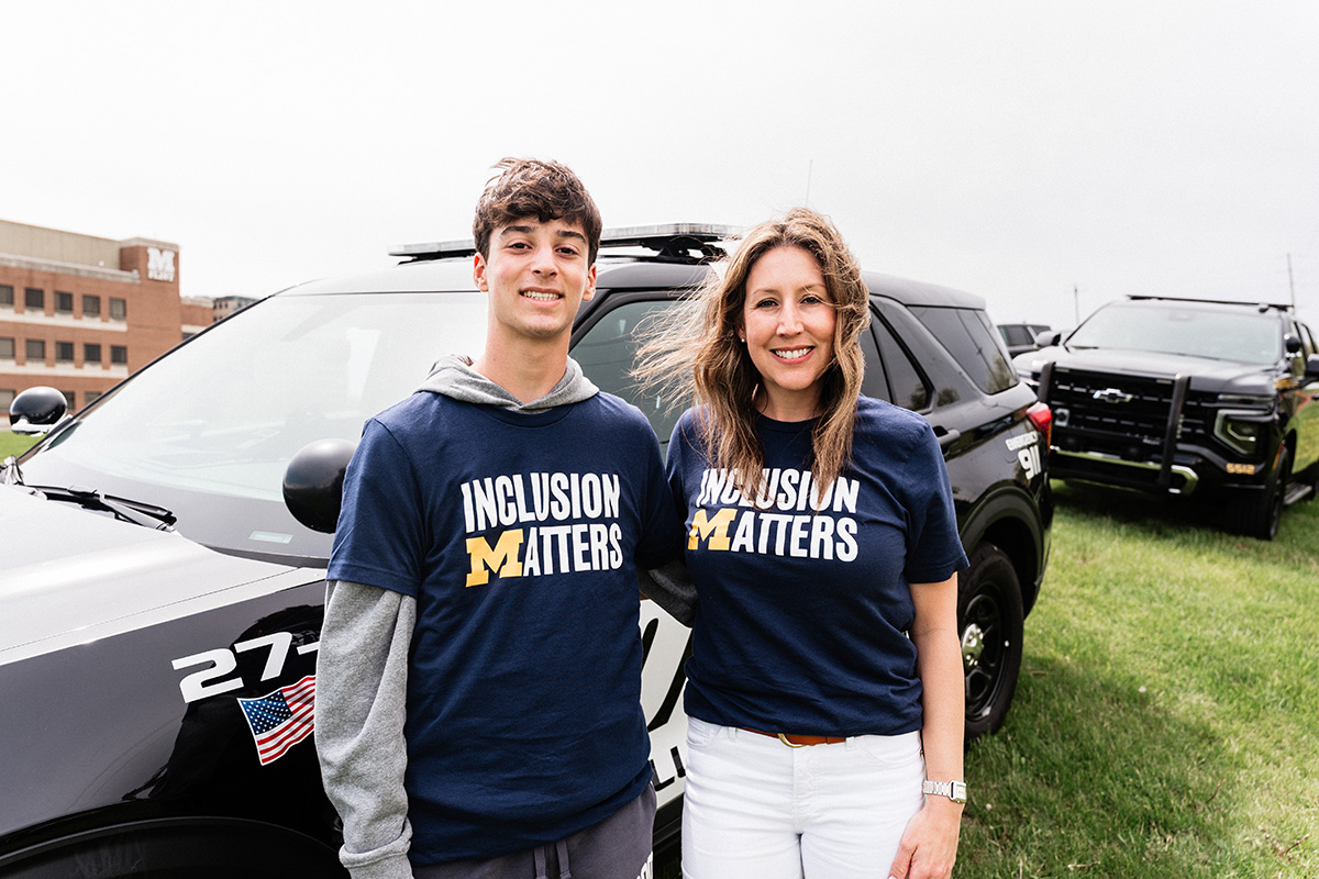 Two people wearing navy blue "Inclusion Matters" t-shirts stand in front of police vehicles on grass.