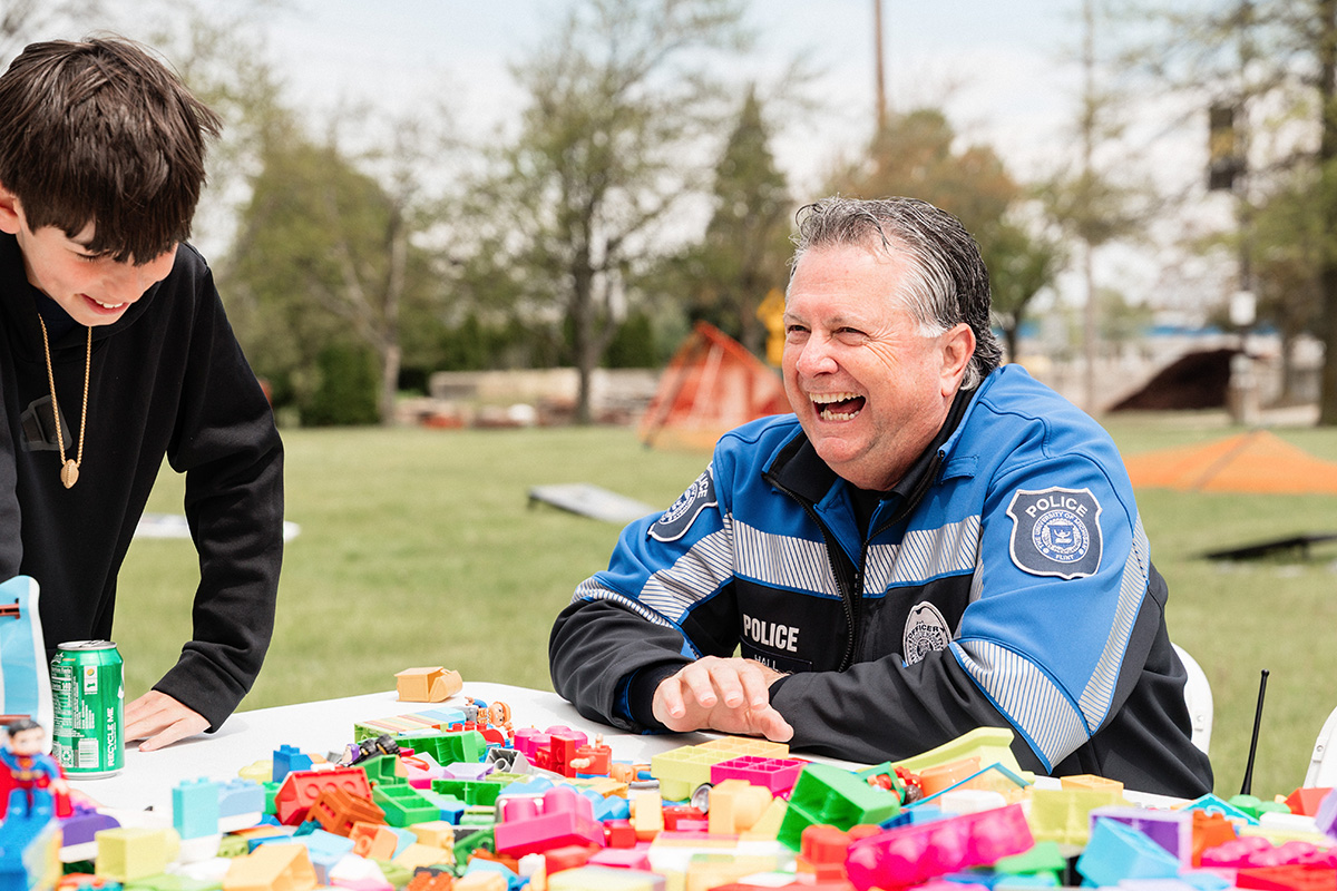 A police officer and a young person interacting at a table covered with colorful building blocks outdoors.