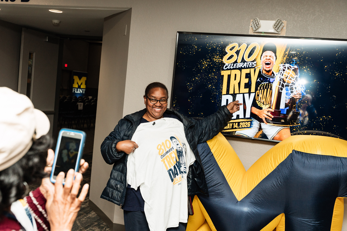 FLINT_Trey Day 2026_0004 Person holding a white "810 Celebrates Trey Day" t-shirt in front of a large screen with the same text and a basketball player holding a trophy.
