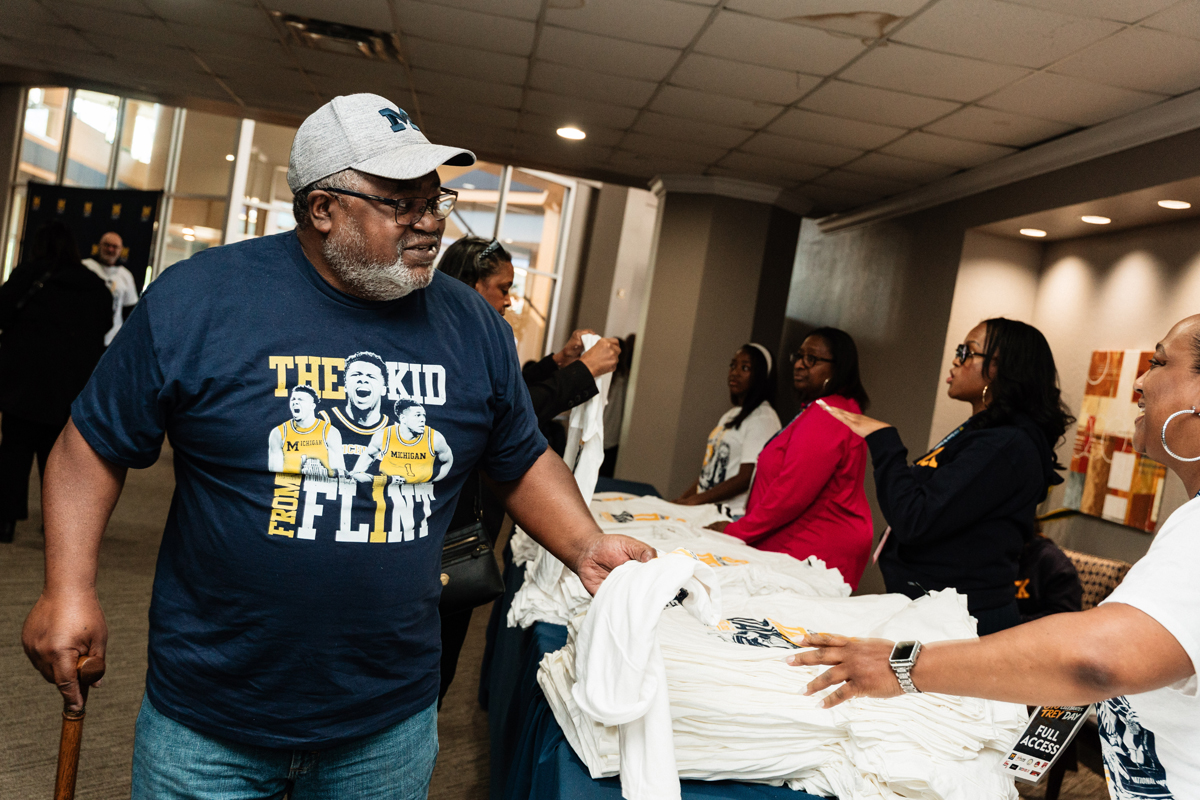 FLINT_Trey Day 2026_0012 Man wearing a navy blue "The Kid From Flint" T-shirt holding a white T-shirt at a table with stacks of white T-shirts and people behind the table.