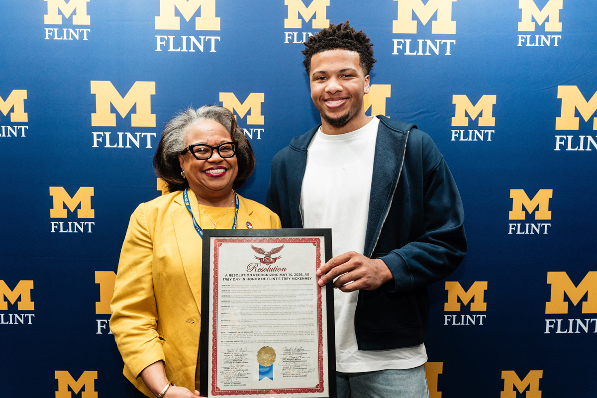 FLINT_Trey Day 2026_0025 Two people holding a framed resolution certificate in front of a blue backdrop with multiple yellow "M Flint" logos.