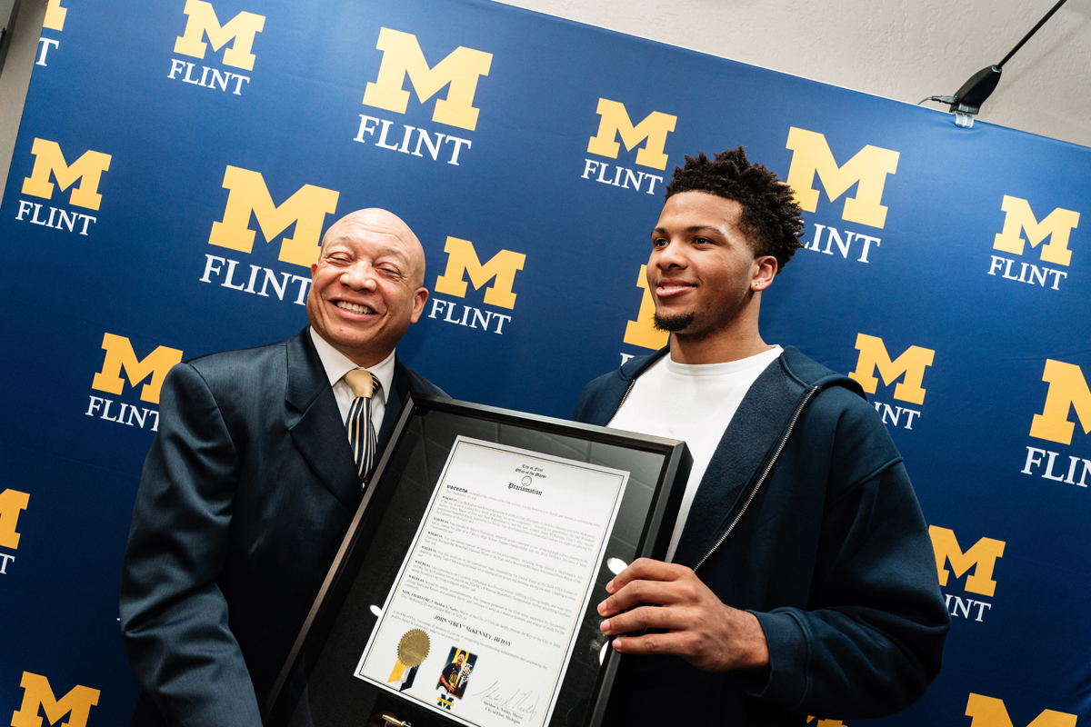 FLINT_Trey Day 2026_0027 Two people holding a framed proclamation in front of a blue backdrop with multiple yellow "M FLINT" logos.