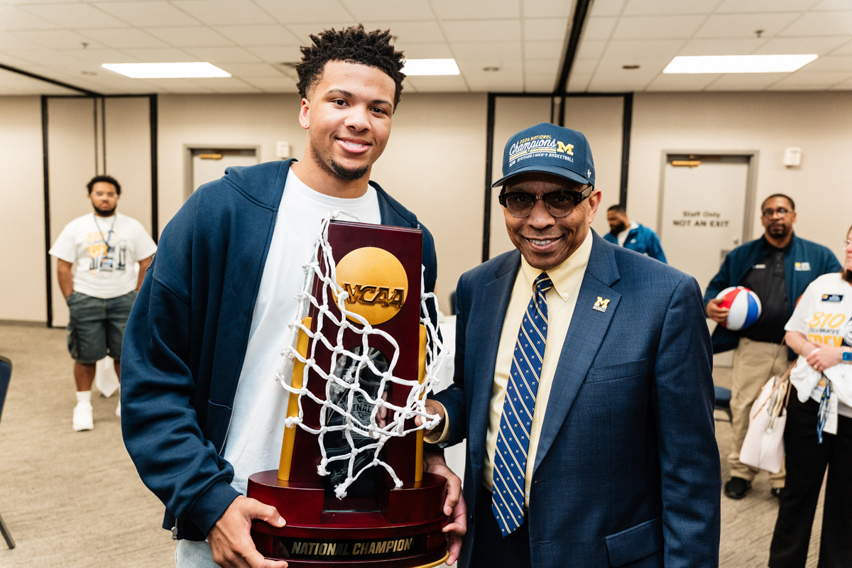 FLINT_Trey Day 2026_0032 Two men holding an NCAA National Champion trophy with a basketball net draped over it in an indoor room.