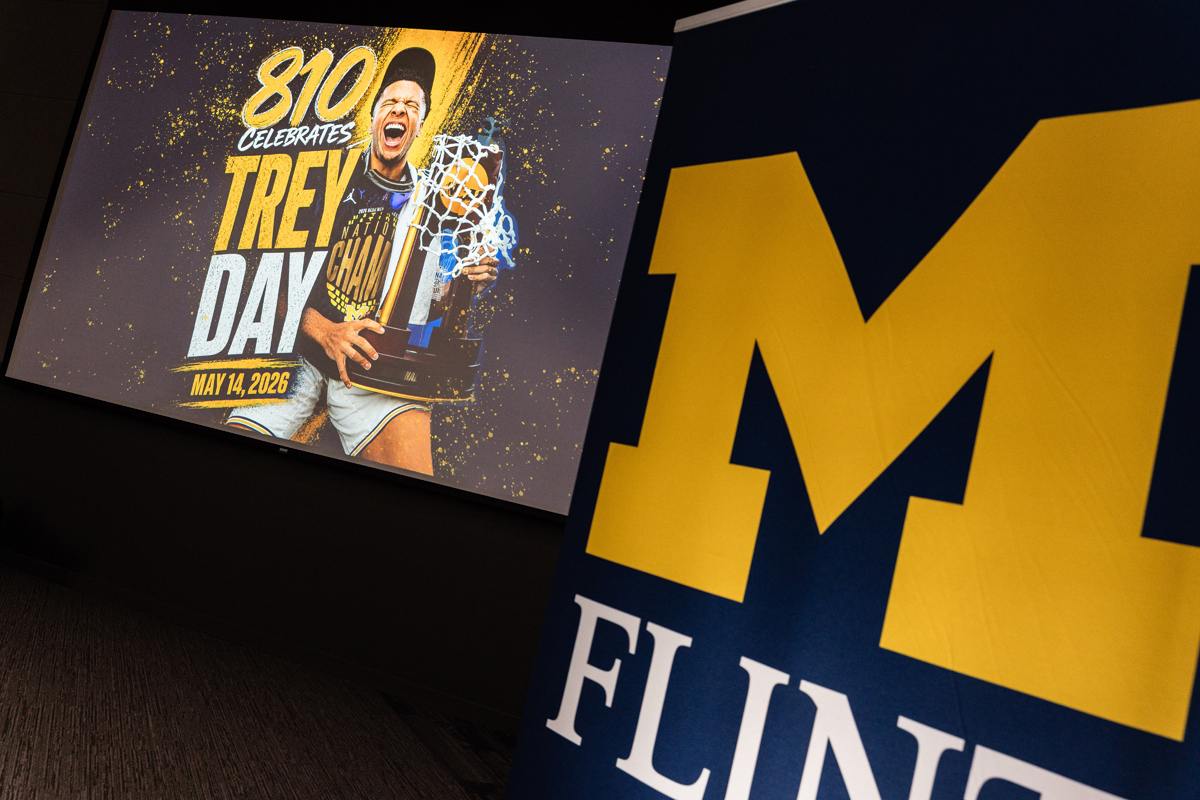 FLINT_Trey Day 2026_0033 Large yellow block "M" with "FLINT" below it on a blue background, next to a screen showing a basketball player holding a trophy with text "810 Celebrates Trev Day May 14, 2026"