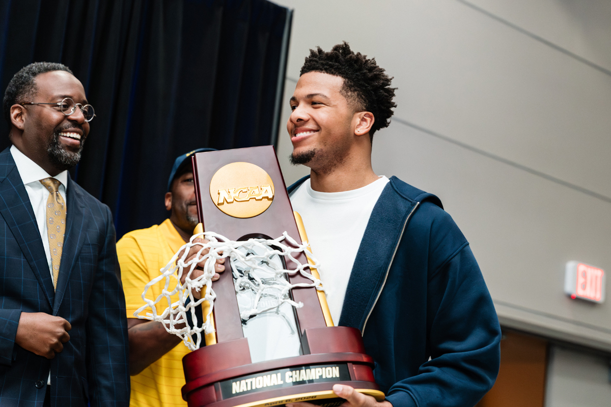 Three people holding an NCAA National Champion trophy with a basketball net attached.