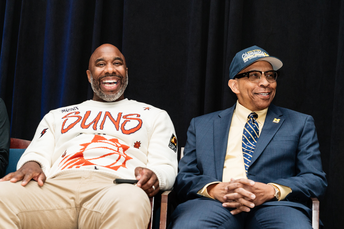 FLINT_Trey Day 2026_0045 Two men sitting side by side; the man on the left wears a white Phoenix Suns sweater, the man on the right wears a blue suit and a 2023 National Champions cap.