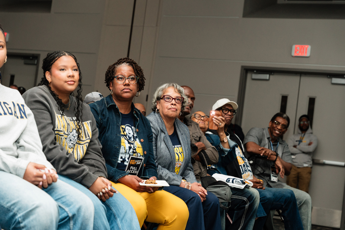 FLINT_Trey Day 2026_0049 A group of people sitting indoors, some wearing University of Michigan apparel, with one person holding a plate of food and another taking a photo with a smartphone.