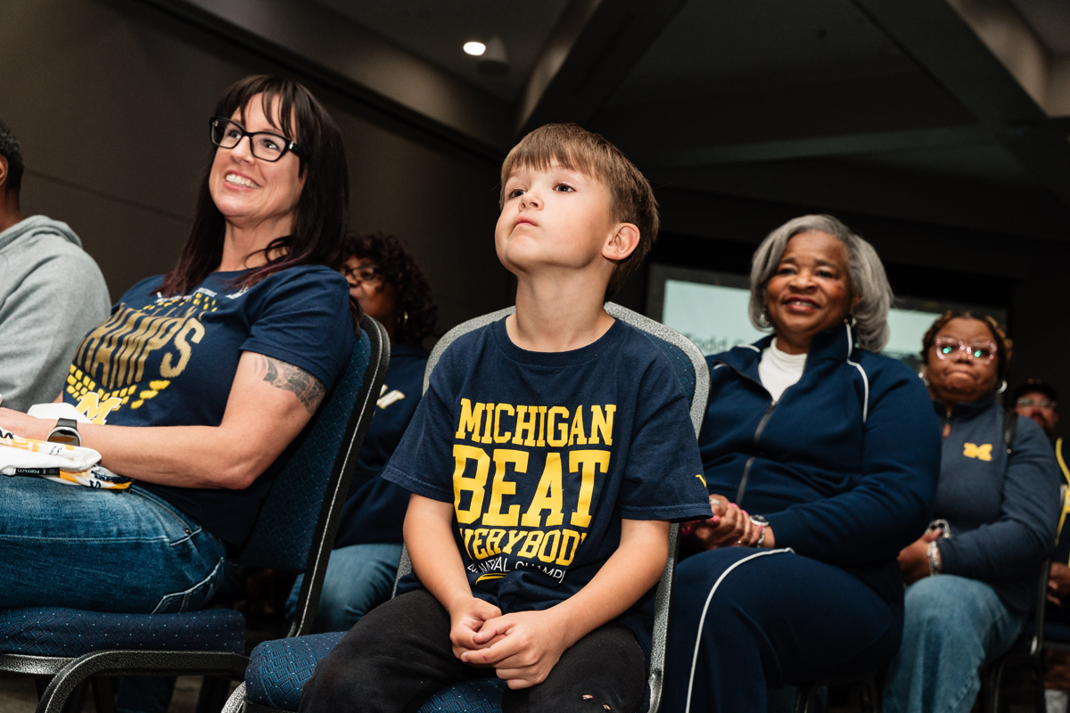 FLINT_Trey Day 2026_0056 A young boy wearing a navy blue "Michigan Beat Everybody" t-shirt sitting among adults in a room.