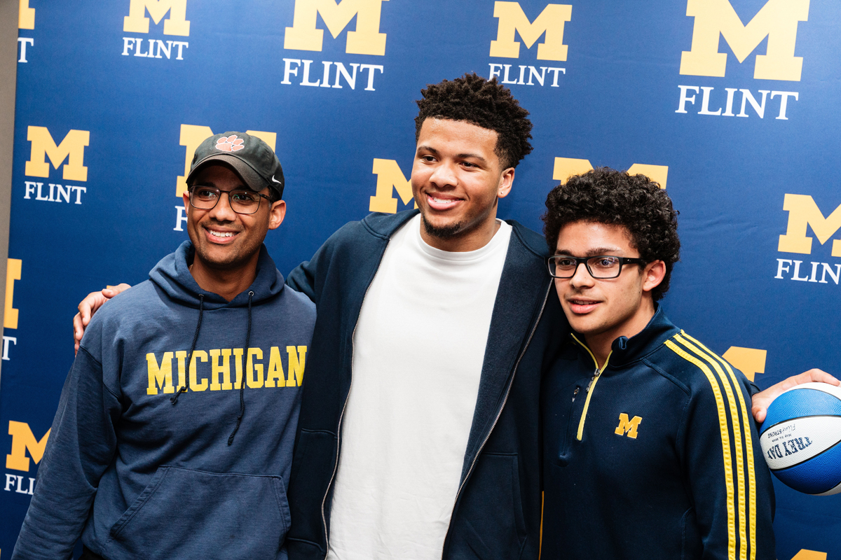 FLINT_Trey Day 2026_0099 Three people standing in front of a blue backdrop with multiple yellow "M FLINT" logos, one holding a blue and white basketball.