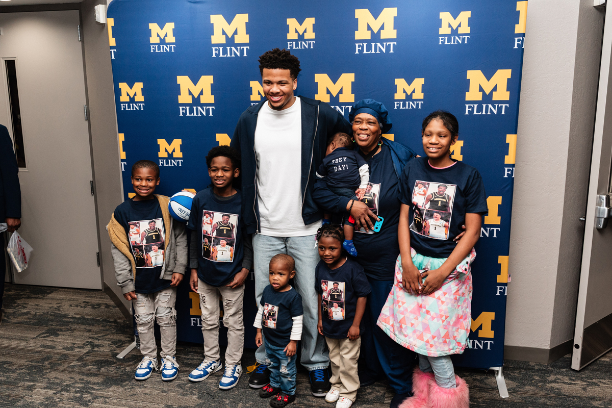 FLINT_Trey Day 2026_0100 Group of people, including children and an adult, standing in front of a blue backdrop with yellow "M Flint" logos, some wearing matching shirts with a basketball player image.