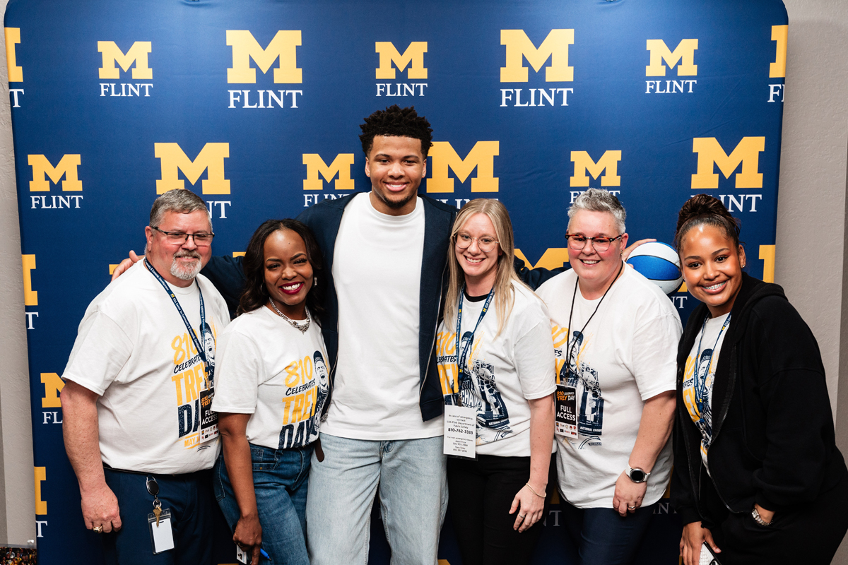 FLINT_Trey Day 2026_0103 Six people posing in front of a blue backdrop with yellow "M Flint" logos, five wearing white event t-shirts.