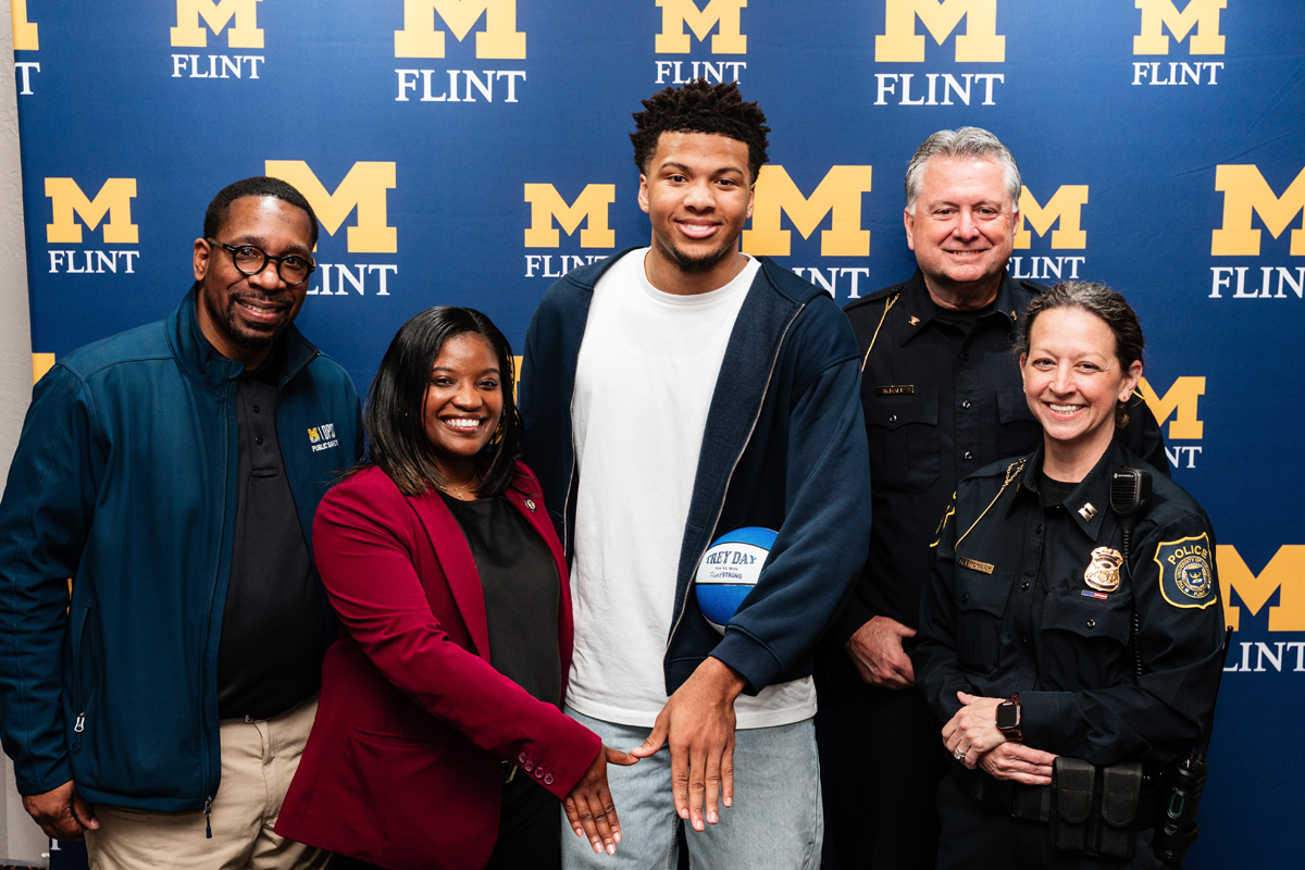 FLINT_Trey Day 2026_0108 Five people standing in front of a blue backdrop with yellow "M FLINT" logos, one holding a basketball.