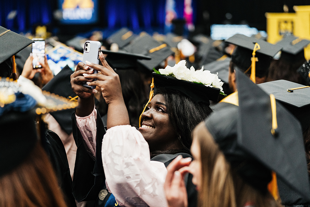 FLINT_Winter Commencement April 2026_0011 A group of graduates wearing black caps and gowns, with one graduate decorated with white flowers on their cap taking a photo with a smartphone.