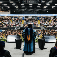 View from behind a speaker in academic regalia addressing a large audience of graduates and attendees in a decorated indoor arena.