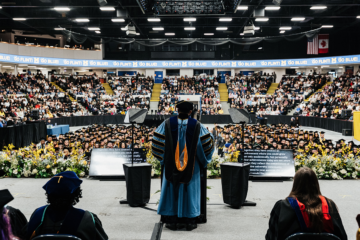 View from behind a speaker in academic regalia addressing a large audience of graduates and attendees in a decorated indoor arena.