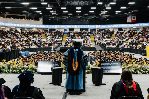 View from behind a speaker in academic regalia addressing a large audience of graduates and attendees in a decorated indoor arena.