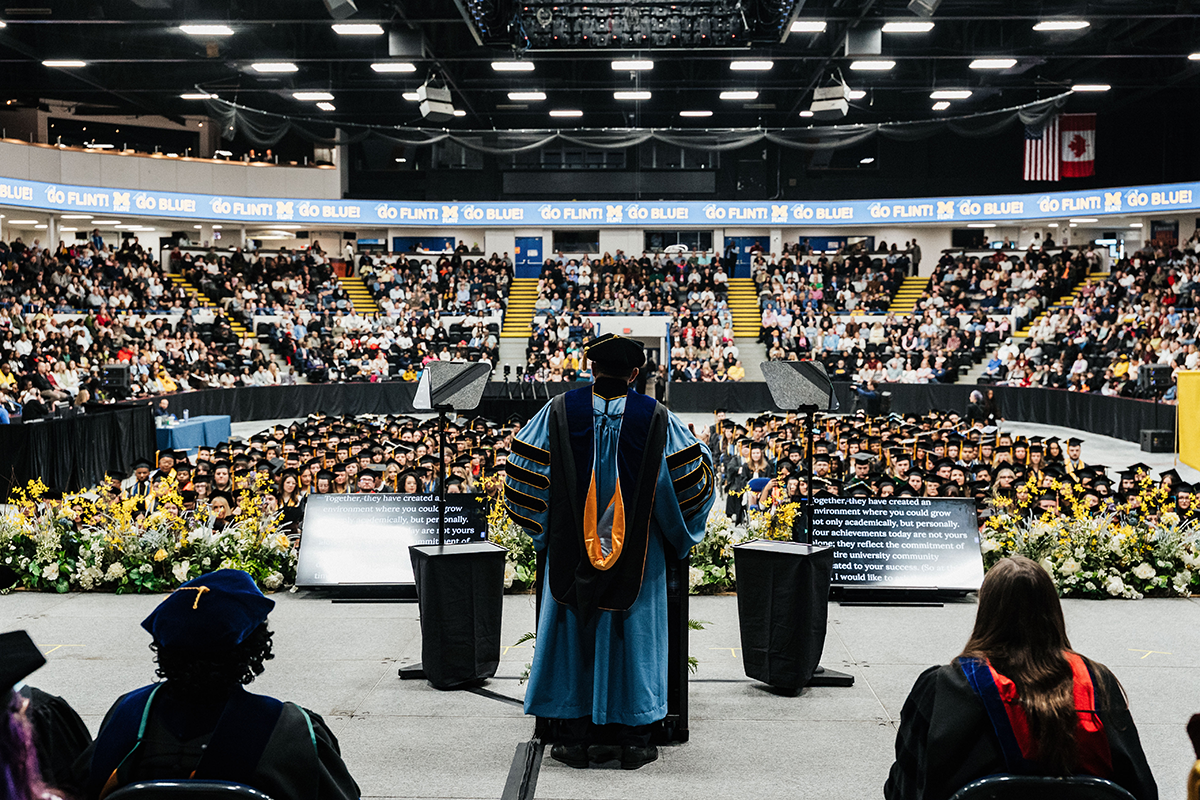 View from behind a speaker in academic regalia addressing a large audience of graduates and attendees in a decorated indoor arena.