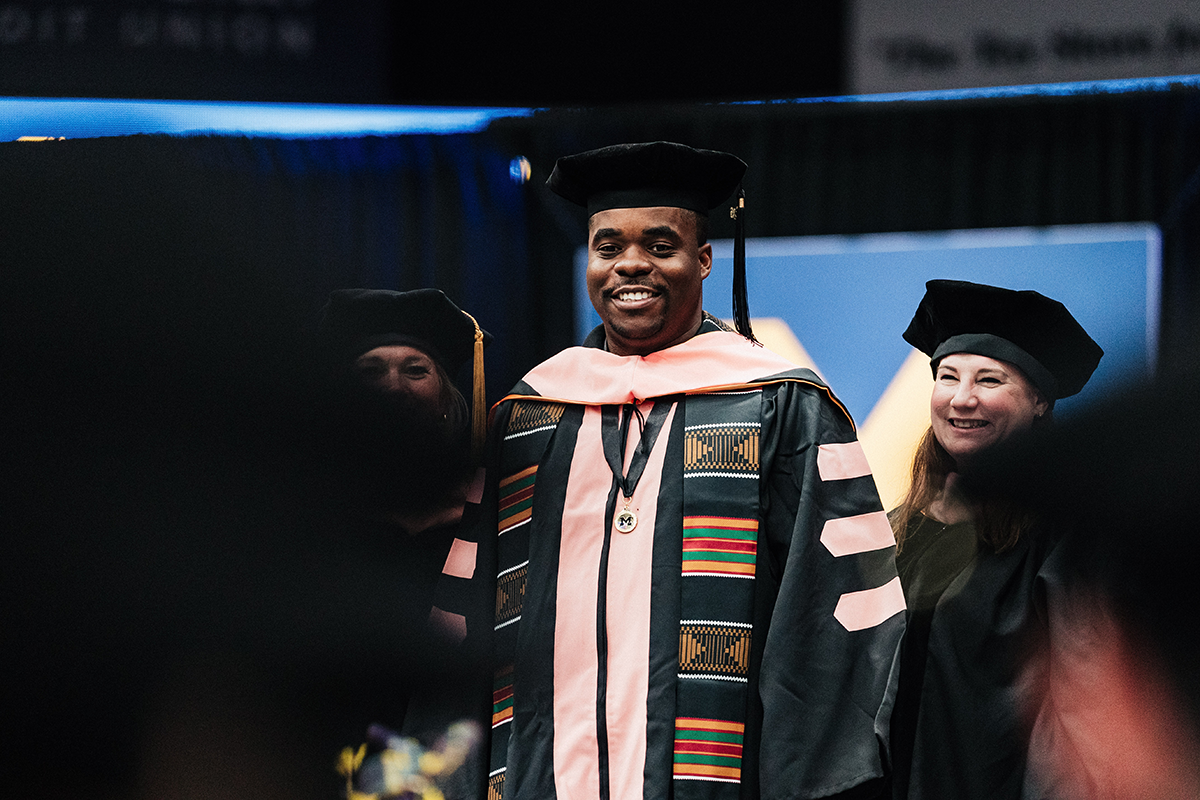 FLINT_Winter Commencement April 2026_0074 A person in academic regalia with a black cap and gown decorated with colorful kente cloth patterns, standing on stage during a graduation ceremony.