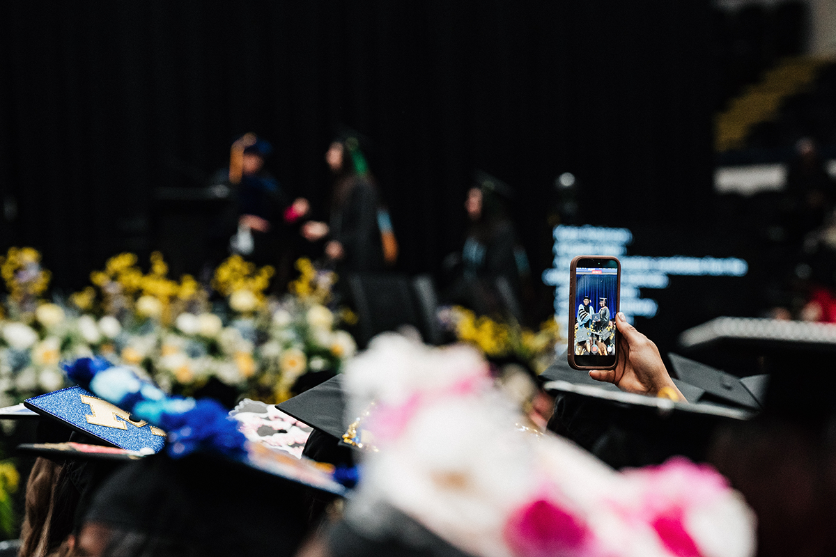 FLINT_Winter Commencement April 2026_0087 A person in graduation attire holding up a phone taking a photo of two graduates on stage.