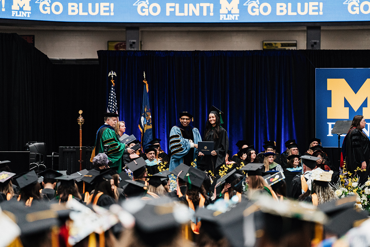 FLINT_Winter Commencement April 2026_0093 Graduates in caps and gowns at a University of Michigan-Flint commencement ceremony receiving diplomas on stage.
