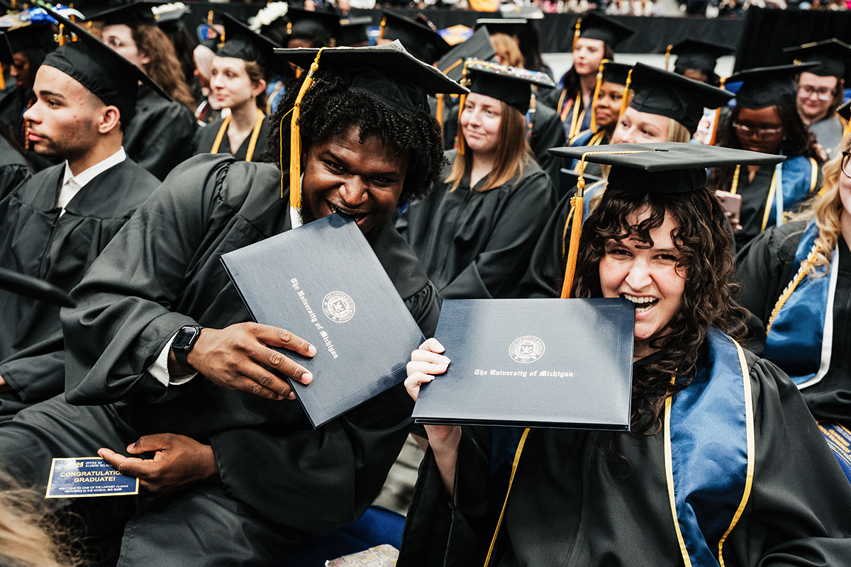FLINT_Winter Commencement April 2026_0137 Two graduates in caps and gowns holding University of Michigan diplomas among a seated crowd at a graduation ceremony.