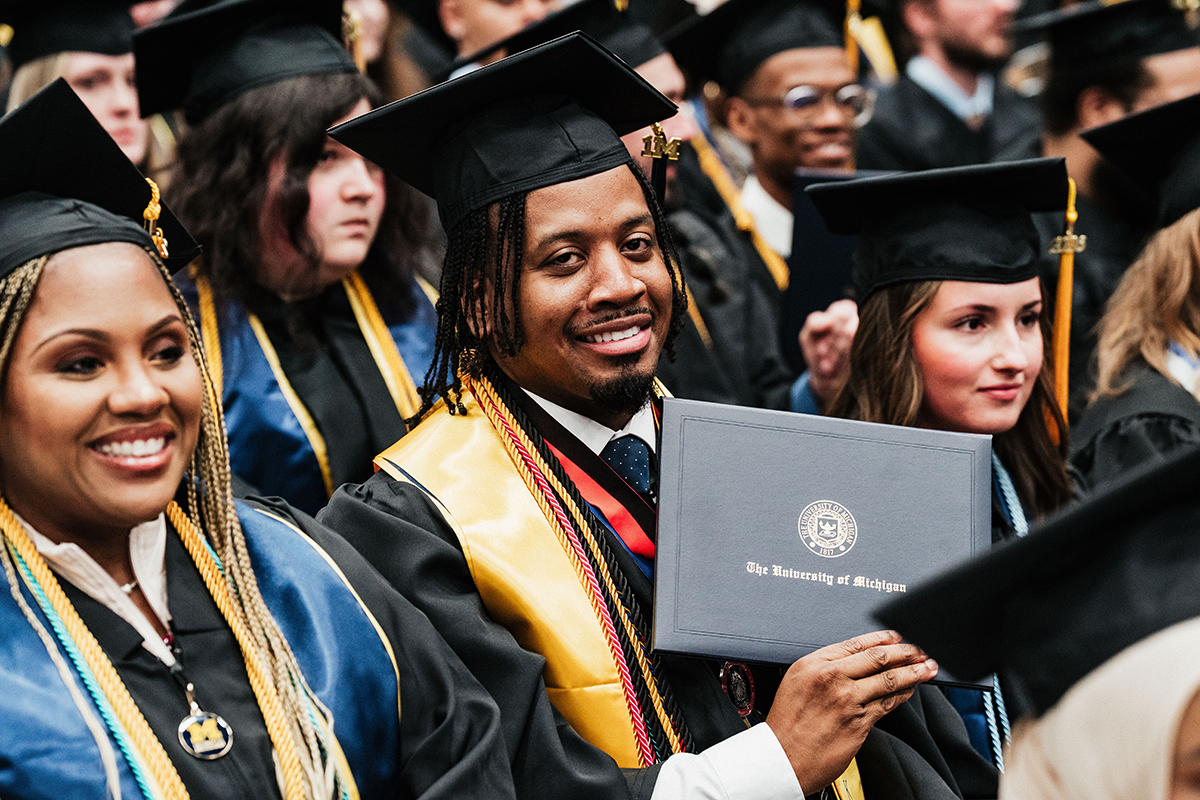 FLINT_Winter Commencement April 2026_0158 A graduate in cap and gown holding a University of Michigan diploma cover during a graduation ceremony.