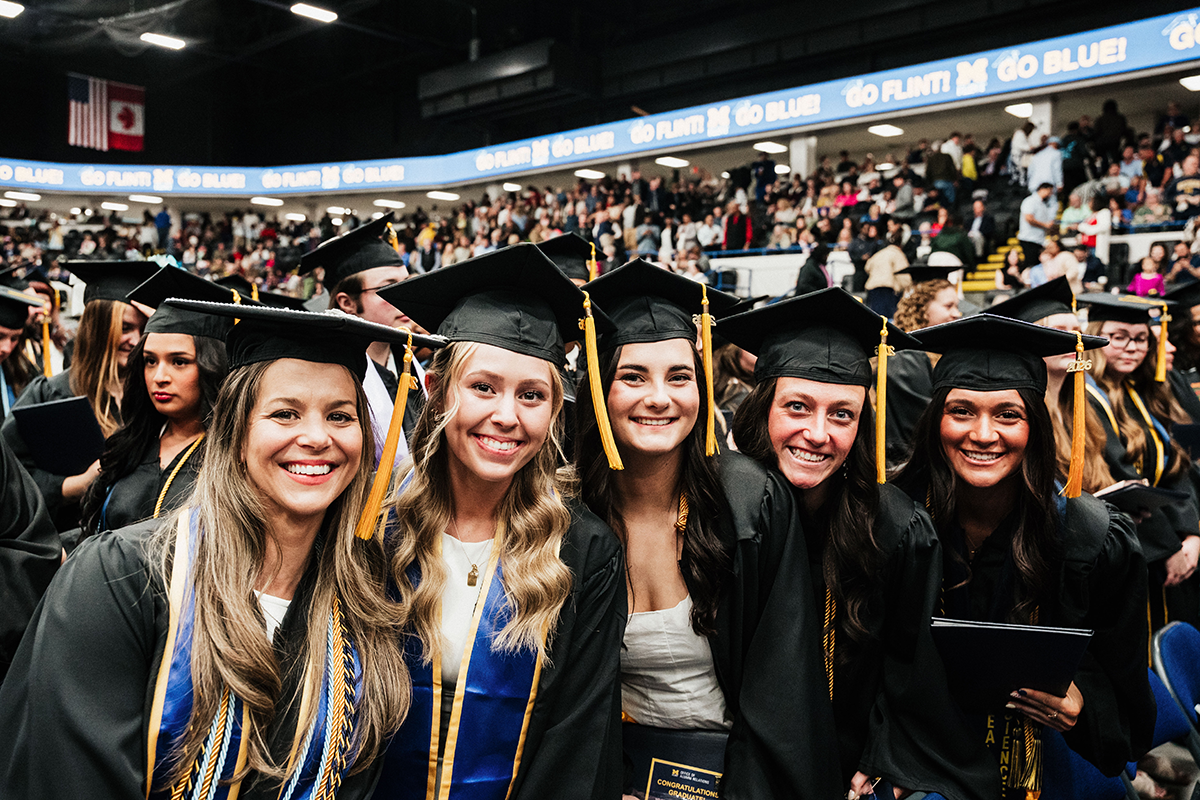 FLINT_Winter Commencement April 2026_0162 Group of graduates in caps and gowns sitting together at a graduation ceremony in an indoor arena.