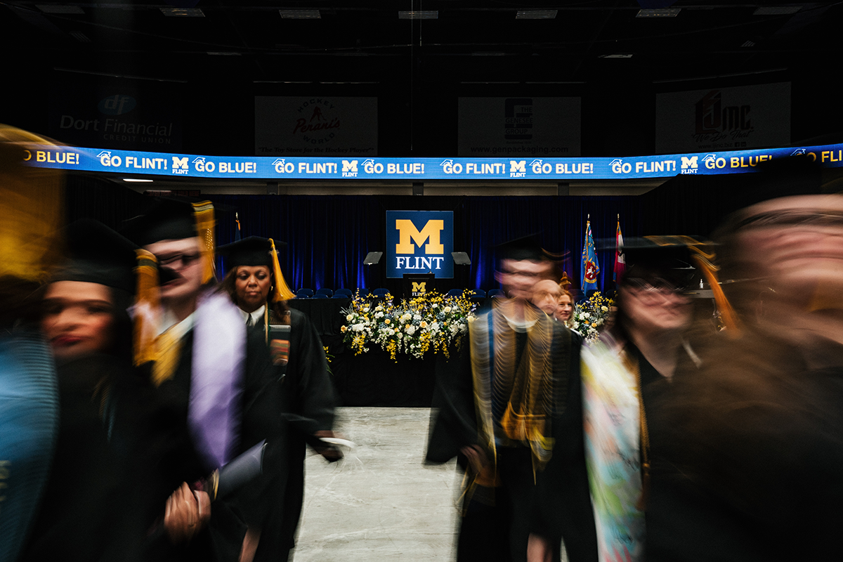 FLINT_Winter Commencement April 2026_0167 Graduates in caps and gowns walking in front of a stage with a University of Michigan-Flint logo and a banner reading "GO FLINT! GO BLUE!"