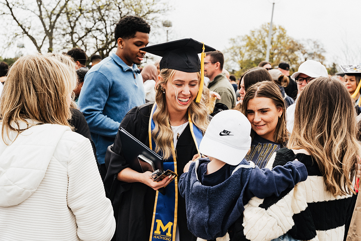FLINT_Winter Commencement April 2026_0179 A graduate in a cap and gown with a University of Michigan Health Science stole is surrounded by family and friends outdoors.