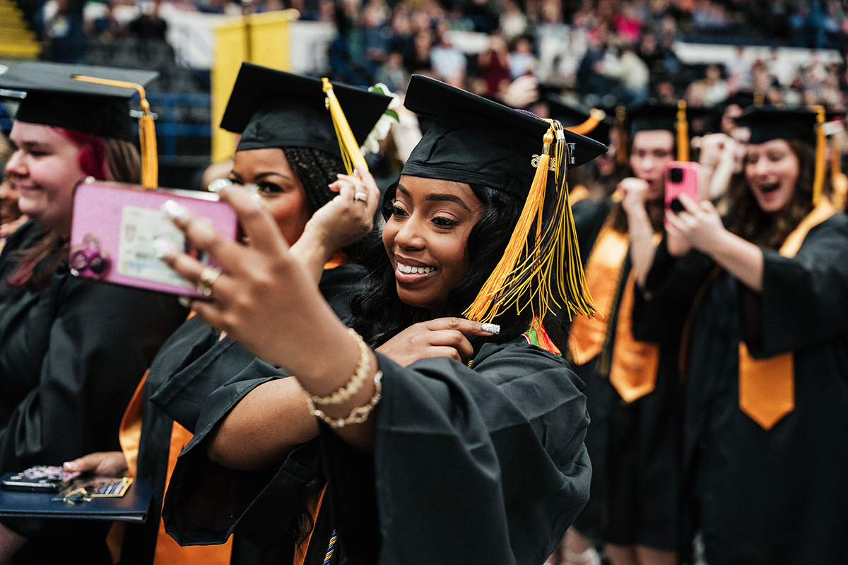FLINT_Winter Commencement April 2026_0194 Group of graduates in caps and gowns taking selfies during a graduation ceremony.