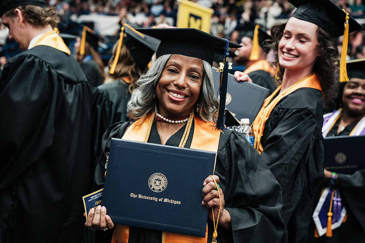FLINT_Winter Commencement April 2026_0198 Graduate in cap and gown holding a University of Michigan diploma at a commencement ceremony.