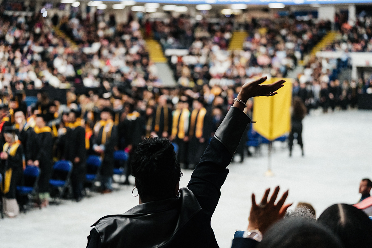 FLINT_Winter Commencement April 2026_0212 Person in a black jacket raising their hand at a graduation ceremony with graduates and audience in the background.