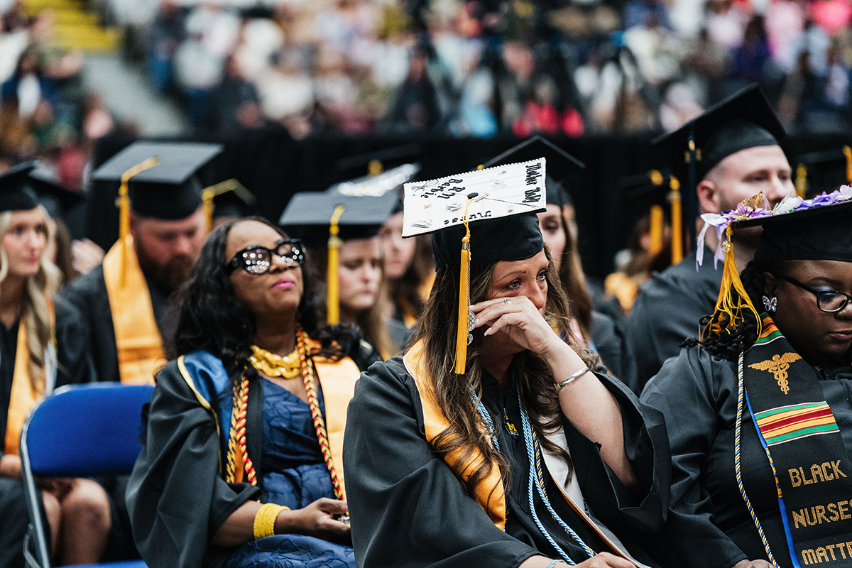 FLINT_Winter Commencement April 2026_0239 Graduates in caps and gowns seated at a graduation ceremony, one wiping tears from her face.