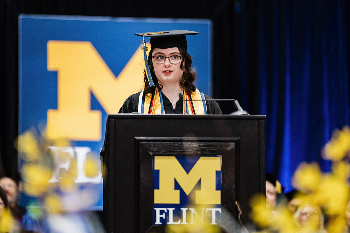 FLINT_Winter Commencement April 2026_0247 A graduate in cap and gown speaking at a podium with a University of Michigan-Flint logo in the background.