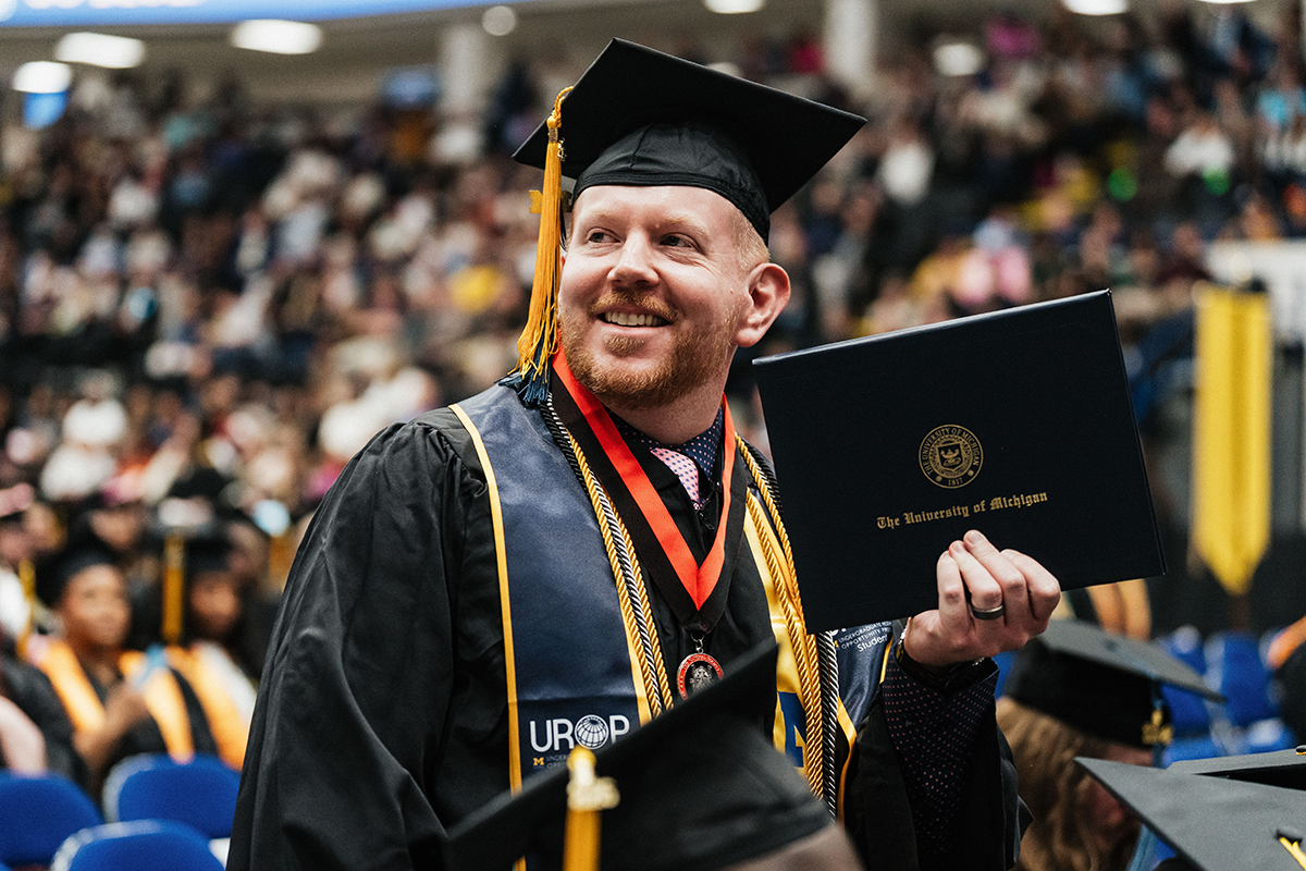 FLINT_Winter Commencement April 2026_0274 Graduate in cap and gown holding a University of Michigan diploma at a commencement ceremony.