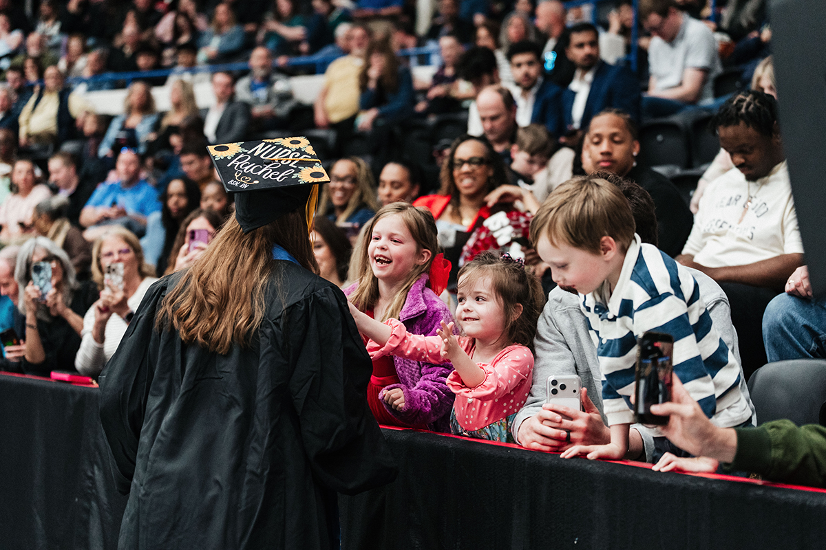 FLINT_Winter Commencement April 2026_0287 Graduate in cap and gown reaching out to children in the audience at a graduation ceremony.