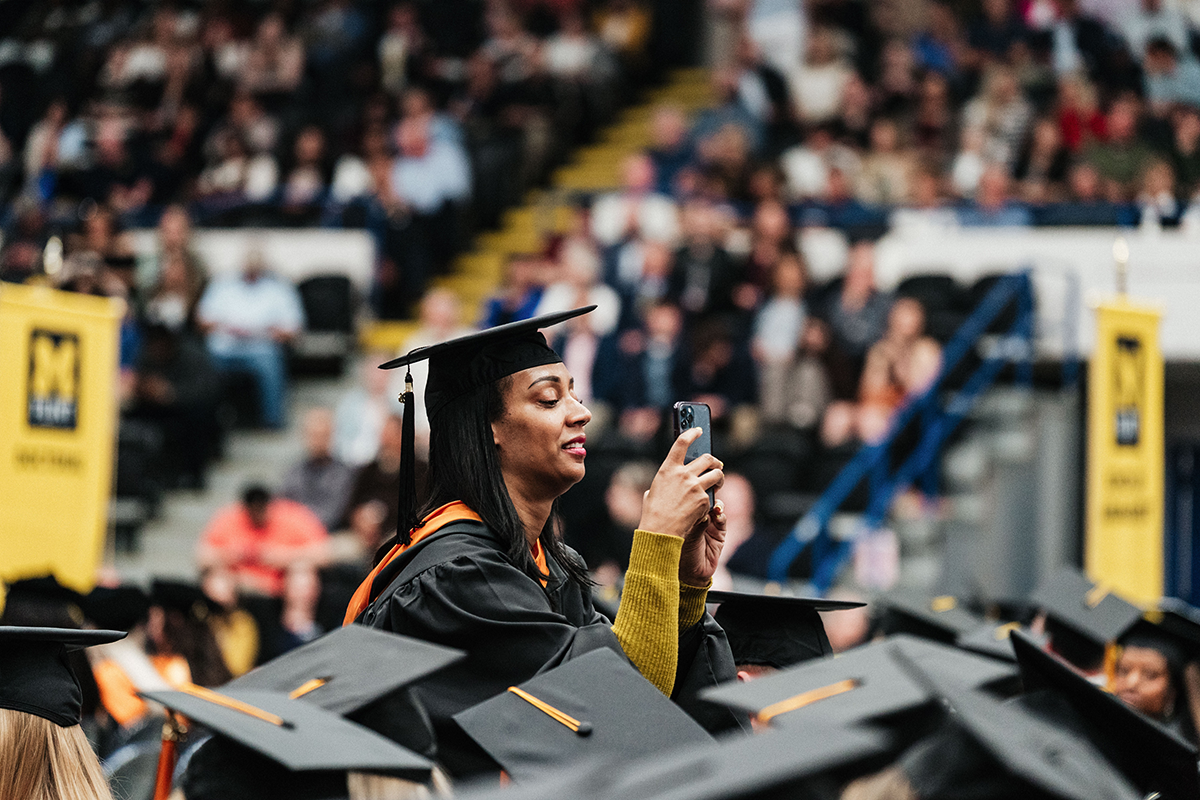 FLINT_Winter Commencement April 2026_0289 A graduate in cap and gown taking a photo with a smartphone during a graduation ceremony.
