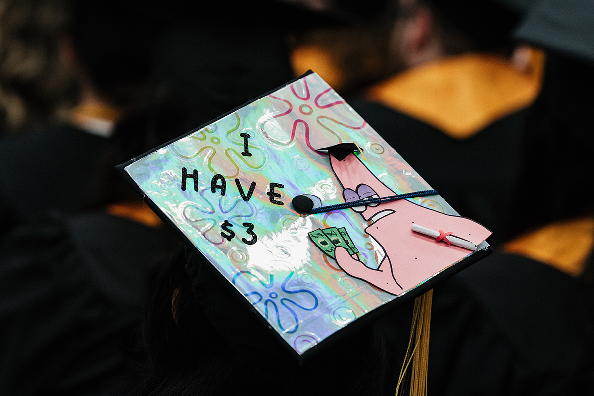 FLINT_Winter Commencement Caps April 2026_0001 Graduation cap decorated with a holographic background, a cartoon character holding money and a diploma, and the text "I HAVE $3".