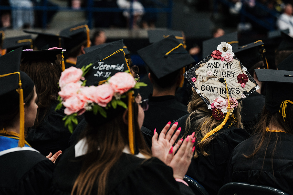 FLINT_Winter Commencement Caps April 2026_0003 Graduates in caps and gowns, one cap decorated with pink flowers, another with "AND MY STORY CONTINUES" and floral decorations.