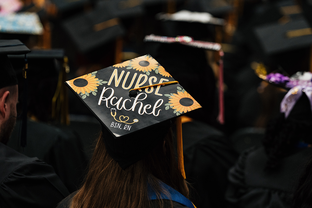 FLINT_Winter Commencement Caps April 2026_0007 Graduation cap decorated with sunflowers and text "Nurse Rachel BSN, RN" worn by a graduate in a crowd.