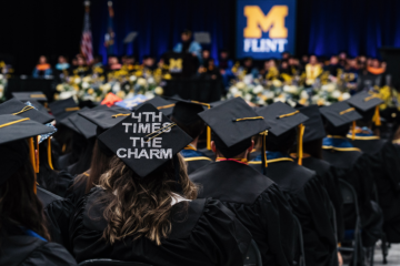 Graduates in caps and gowns at a University of Michigan-Flint graduation ceremony, one cap decorated with "4TH TIMES THE CHARM."