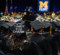 Graduates in caps and gowns at a University of Michigan-Flint graduation ceremony, one cap decorated with "4TH TIMES THE CHARM."