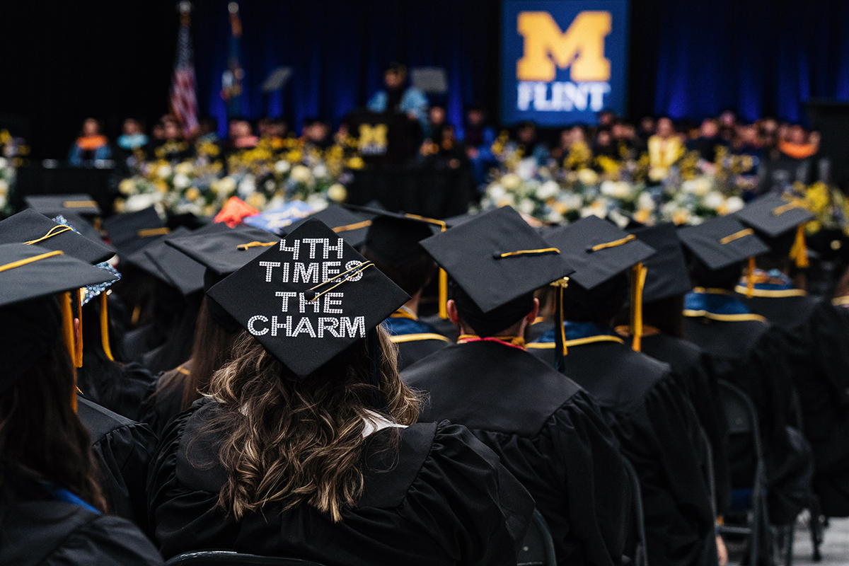 Graduates in caps and gowns at a University of Michigan-Flint graduation ceremony, one cap decorated with "4TH TIMES THE CHARM."