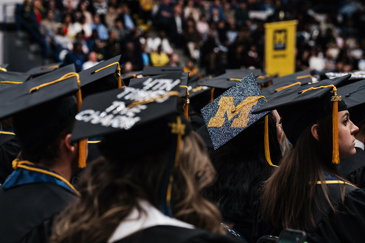 FLINT_Winter Commencement Caps April 2026_0012 Graduates wearing caps and gowns at a university commencement ceremony, one cap decorated with a large glittery gold and blue "M".