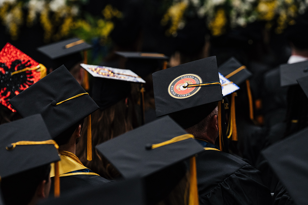 FLINT_Winter Commencement Caps April 2026_0014 Graduates wearing black caps and gowns, one cap decorated with a United States Marine Corps emblem.