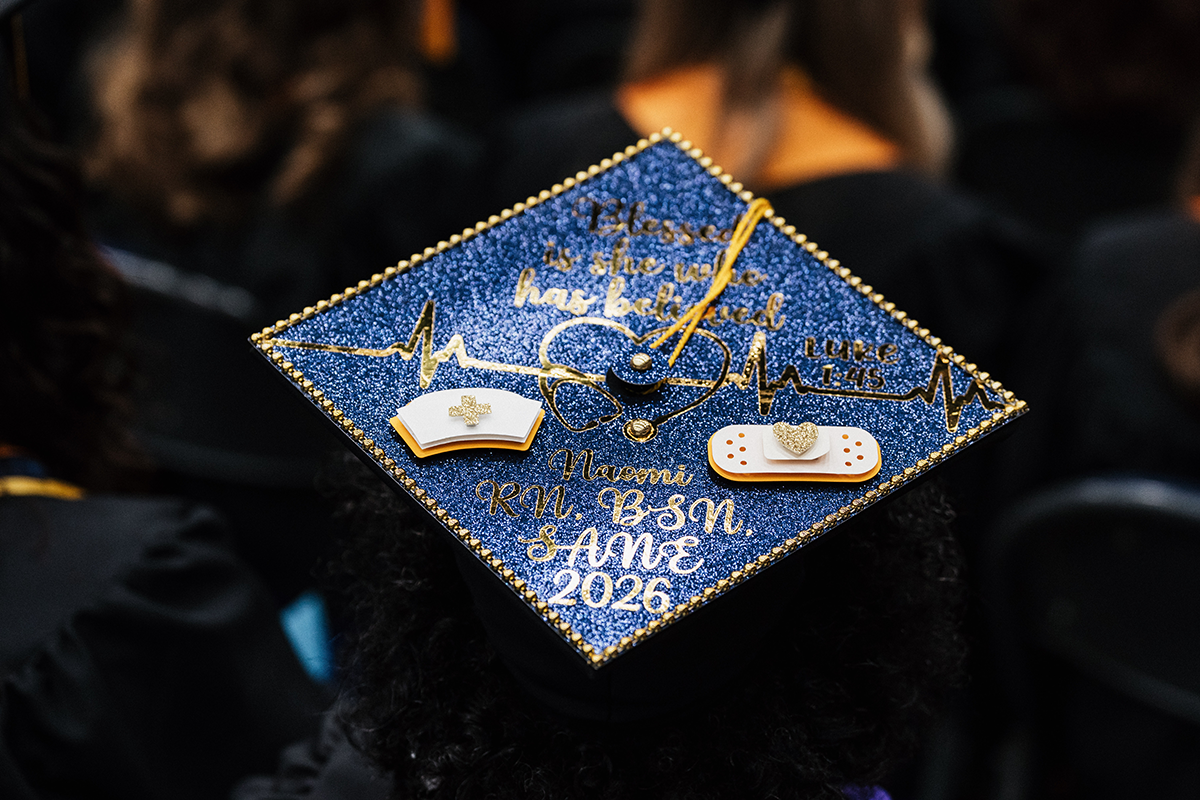 FLINT_Winter Commencement Caps April 2026_0021 Graduation cap decorated with blue glitter, gold heartbeat line, nurse hat, bandage, and text reading "Blessed is she who has believed Luke 1:45 Naomi RN, BSN, SANE 2026".