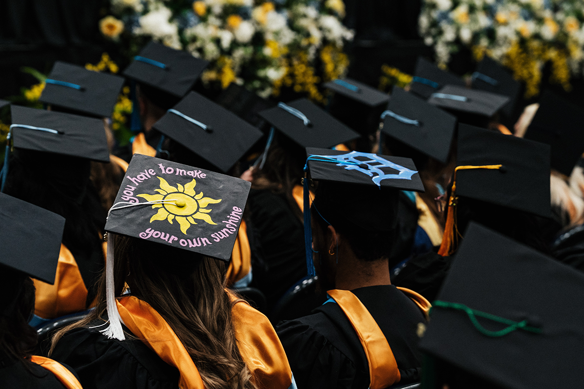 FLINT_Winter Commencement Caps April 2026_0042 Graduates wearing caps and gowns, one cap decorated with a yellow sun and the phrase "you have to make your own sunshine."